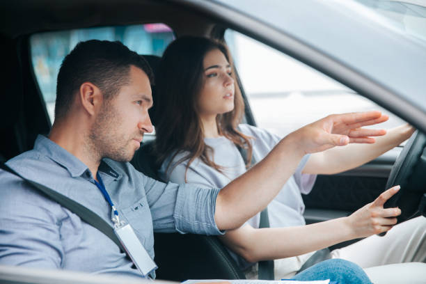 Male instructor showing something by his hand on the road and giving advice. Attractive woman listening her teacher, looking ahead with concentration and holding wheel